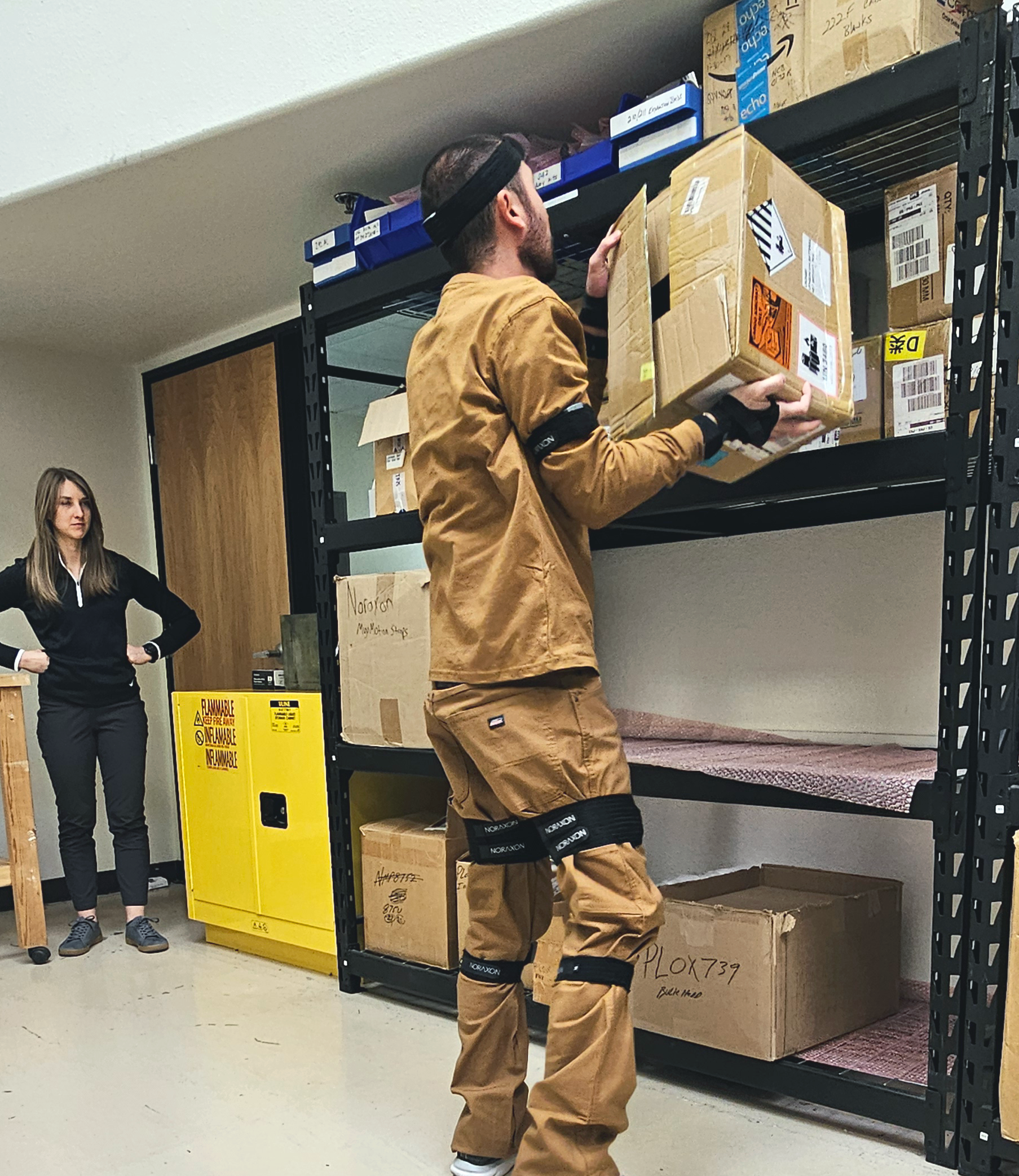 Devon Lifting A man wearing a brown work uniform and Noraxon Ultium Motion capture sensors on his head, arms, wrists, and legs lifts a cardboard box onto a high shelf in a storage area. His posture and movement are being observed by a woman in a black long-sleeve top and dark pants, standing nearby with her hands on her hips. The setting includes industrial shelving filled with labeled boxes, a yellow flammable materials cabinet, and a wooden workstation, indicating an ergonomics or biomechanics research environment.