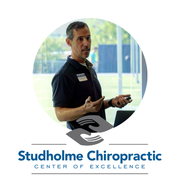 Man giving a presentation while holding a remote, with the logo and text ‘Studholme Chiropractic Center of Excellence’ displayed below.