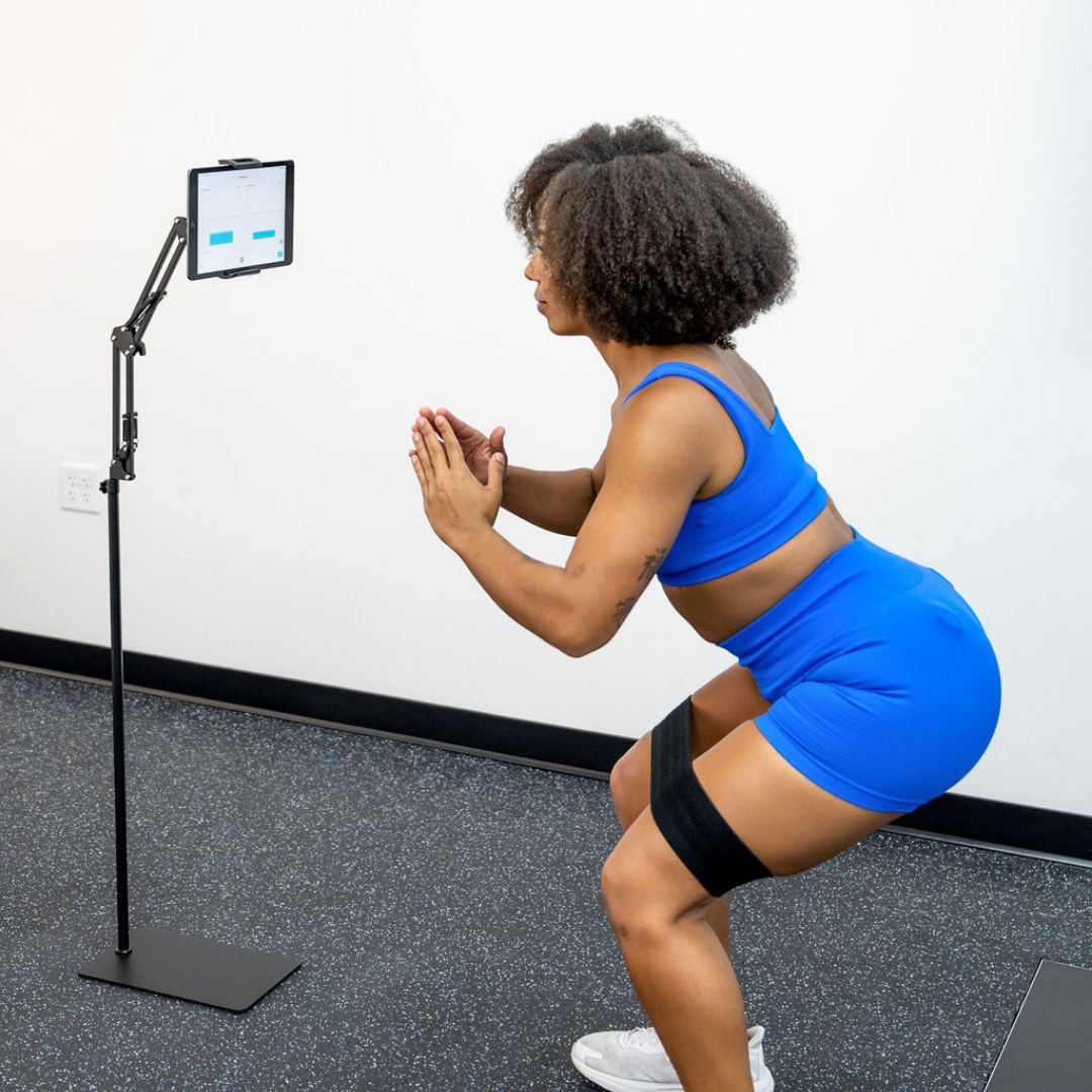 Person performing a squat exercise with a resistance band around the thighs while viewing real-time muscle activity feedback on a tablet using the Noraxon Biofeedback App.