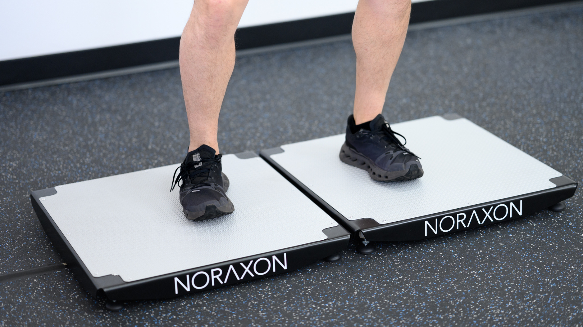 A person stands with one foot on each of two Noraxon force plates, wearing black athletic shoes, demonstrating a stance assessment on a black speckled gym floor. The image focuses on the lower legs and feet, with the Noraxon branding clearly visible on the front of each plate.