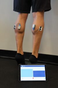 Biofeedback Ultium EMG Wireless EMG sensors attached to both calf muscles during a heel-raise exercise, with live muscle activity displayed on a tablet.