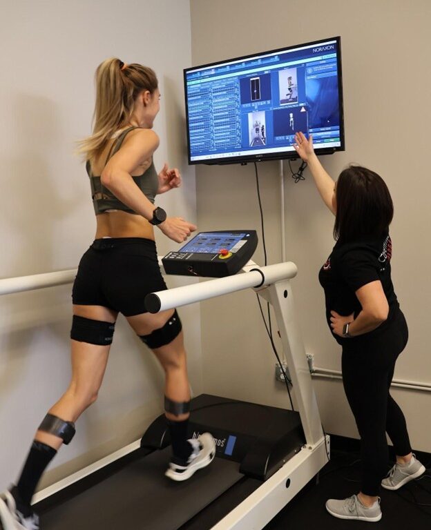 CompEdge Running Athlete runs on a treadmill wearing motion sensors while a clinician reviews real-time movement analysis data on a Noraxon display.